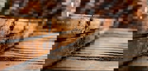 logo Närbild på staplade, väderbitna träbrädor med tydlig textur och spår av åldrande. Fotot är taget i låg vinkel med skärpa på brädornas kant och suddig bakgrund.