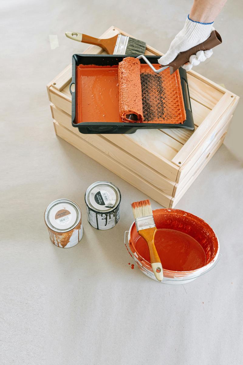 Närbild på målarutrustning med roller, penslar och färgburkar i orange färg, placerade på ett skyddat underlag.