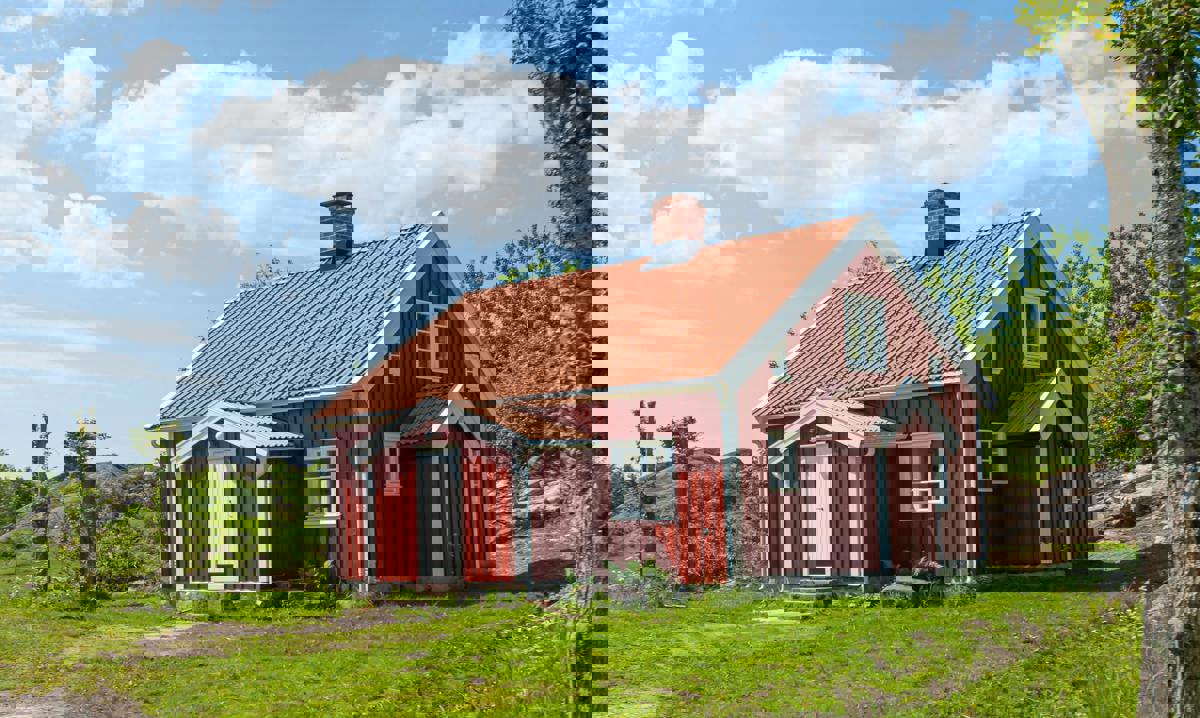 Rött trähus med vita knutar och tegeltak, omgivet av grönska och blå himmel en solig dag.