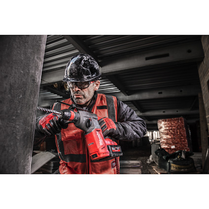 Person i röd arbetsjacka och skyddshjälm använder en röd borrmaskin i industriell miljö.