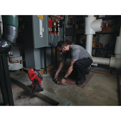 Person i arbetskläder undersöker maskindelar i ett rum med rör, en röd verktygslampa placerad nära.