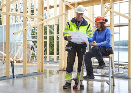 Två personer i byggkläder granskar ritningar på en byggarbetsplats med trästomme i bakgrunden.