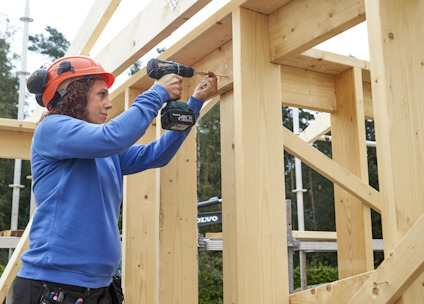 Person i blå tröja och orange hjälm använder borrmaskin på träram vid byggarbetsplats.