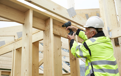 Person i skyddshjälm och reflexjacka använder borrmaskin på träkonstruktion.