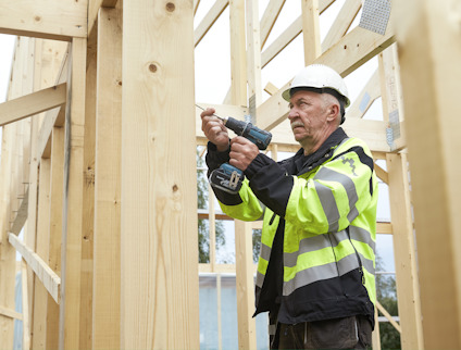 Man i skyddshjälm och reflexjacka använder en borrmaskin på trästomme.