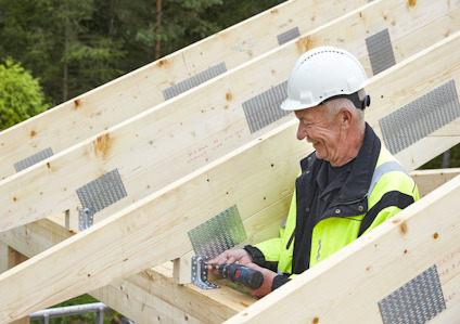 Äldre man i skyddshjälm och gul jacka använder en skruvdragare på träkonstruktion.