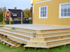 Trädäck byggt med trätrall, intill gult hus med vita fönster och röd byggnad i bakgrunden.