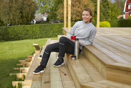 Kvinna i grå tröja sitter på en trätrappa, håller en termos och mugg. Gräsmatta och hus i bakgrunden.