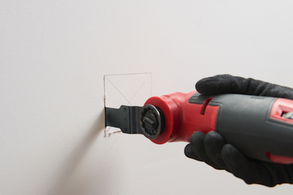 Hand i svart handske använder en elektrisk multiverktygssåg för att skära en kvadratisk form i en vit vägg.