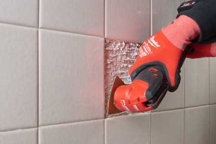 En person använder ett elektriskt verktyg med rött handtag för att skära kakelbeläggning på en vägg.