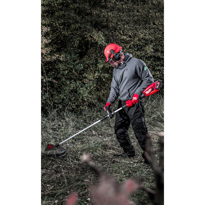 Person med skyddsutrustning använder en grästrimmer i en utomhusmiljö med gräs och buskar.
