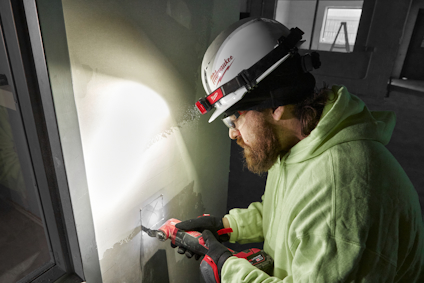 Person i hjälm och skyddsglasögon håller en elektrisk handverktyg, arbetar på en vägg i ett byggprojekt.