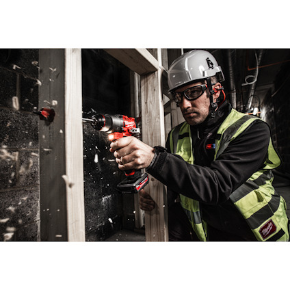 Person i skyddsutrustning använder en borrmaskin på en träregel; spån flyger.