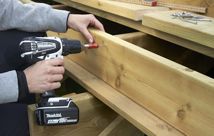 Person använder en borrmaskin för att skruva i en träbalk; skruvar och måttstock syns i bakgrunden.