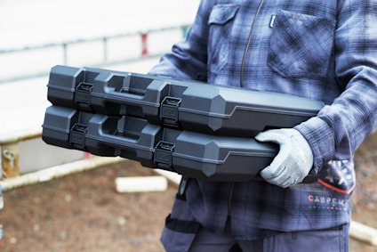 Person bär två svarta verktygslådor med lås, iklädd blå- och vitrutig skjorta samt grå handskar.