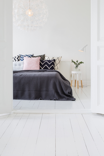 Säng med svarta och rosa kuddar, vit golvlampa, ljust träbord och dekorativ taklampa i vitt rum.