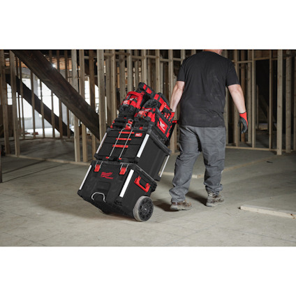 Person som drar en stapel med röda och svarta verktygslådor på hjul i en byggarbetsplats.