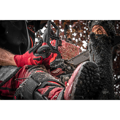 Person använder motorsåg och bär röda skyddskläder, inklusive handskar, i arbete med trädfällning.