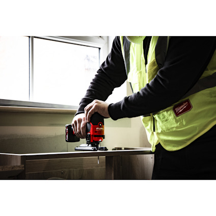 Person i säkerhetsväst använder en röd elektrisk sticksåg på en träyta nära ett fönster.