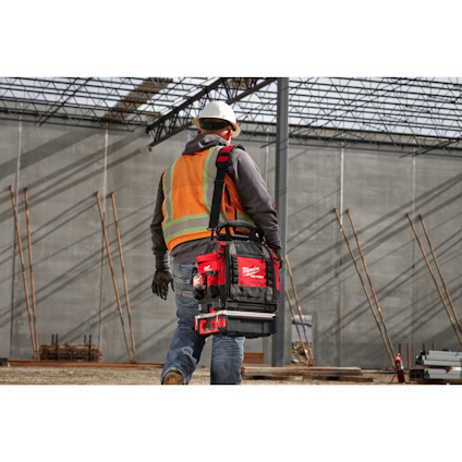 Person i arbetskläder bär en verktygsväska och går mot en byggarbetsplats med betongväggar och stålbjälkar.