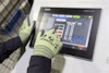 Händer med gröna handskar använder en pekskärm med olika symboler och knappar. Skärmen visar "SMARTC HMI".