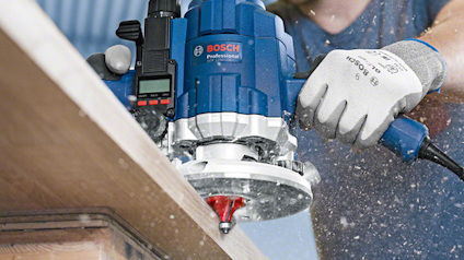 En hand håller i en blå Bosch Professional fräs, som bearbetar en kant av trä. Brus och träspån syns i luften.