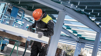 Person med orange hjälm och arbetskläder skär isoleringsmaterial i fabrikslokal.