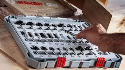 En hand väljer en borr från ett set med Bosch-borrar i en grå verktygslåda på en arbetsbänk.