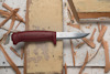Kniv med rött handtag och stålblad på en träyta med träflisor runtomkring.